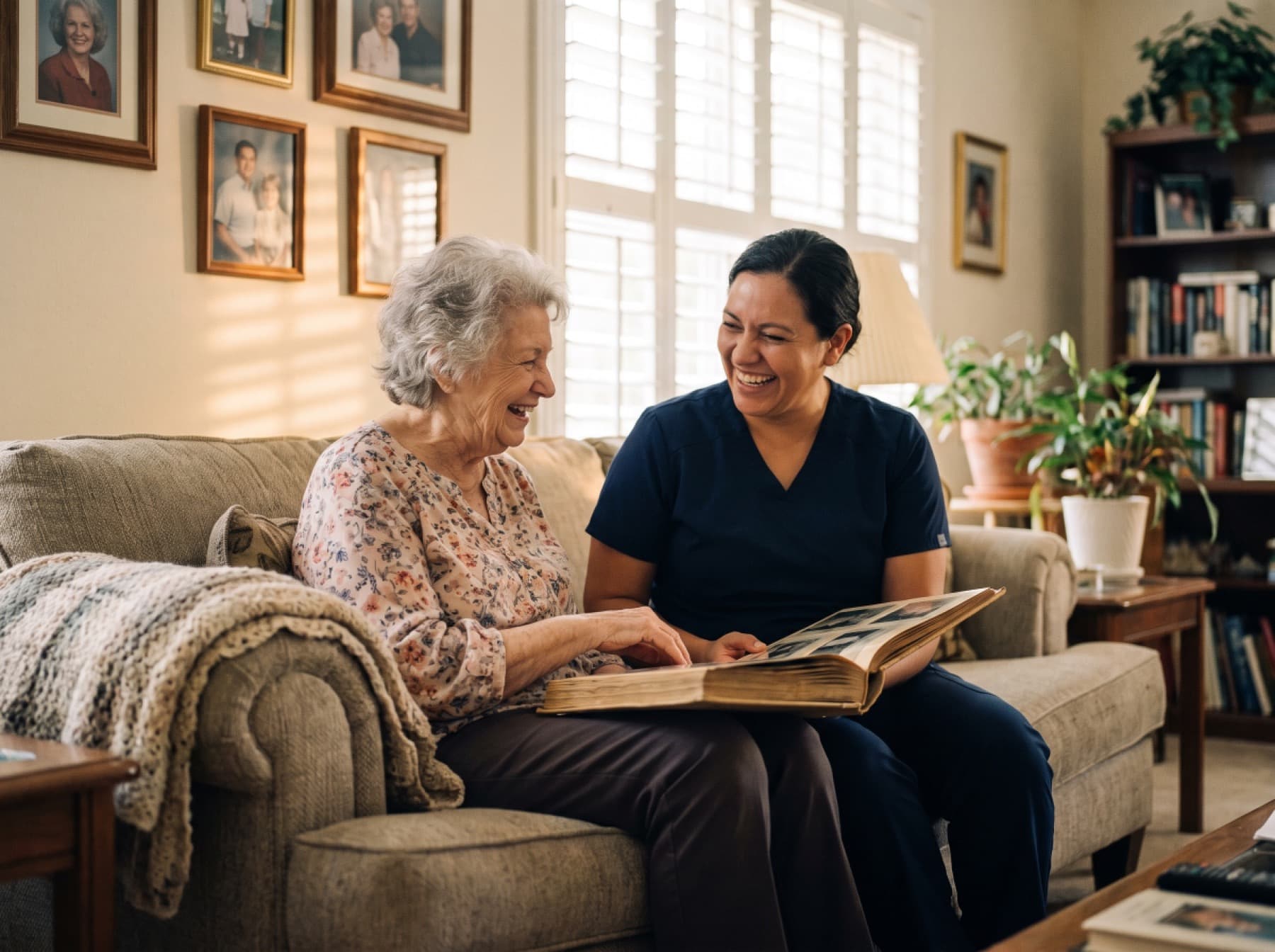 A caregiver in navy scrubs and an older woman laughing together over an open photo album on the sofa.