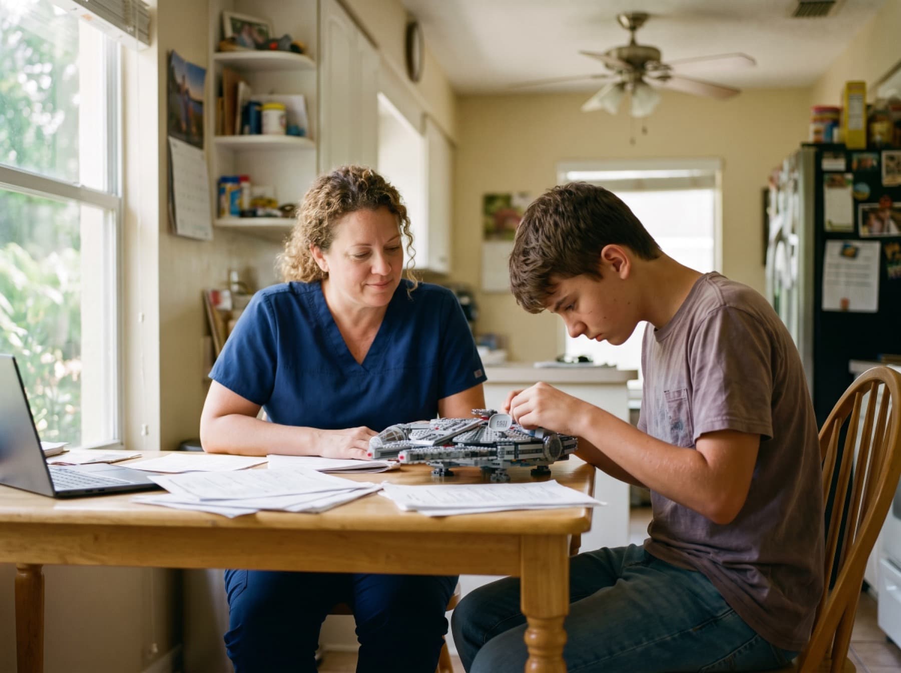 A caregiver in navy scrubs sitting at a kitchen table beside a teenage boy, the two of them working on a model together.
