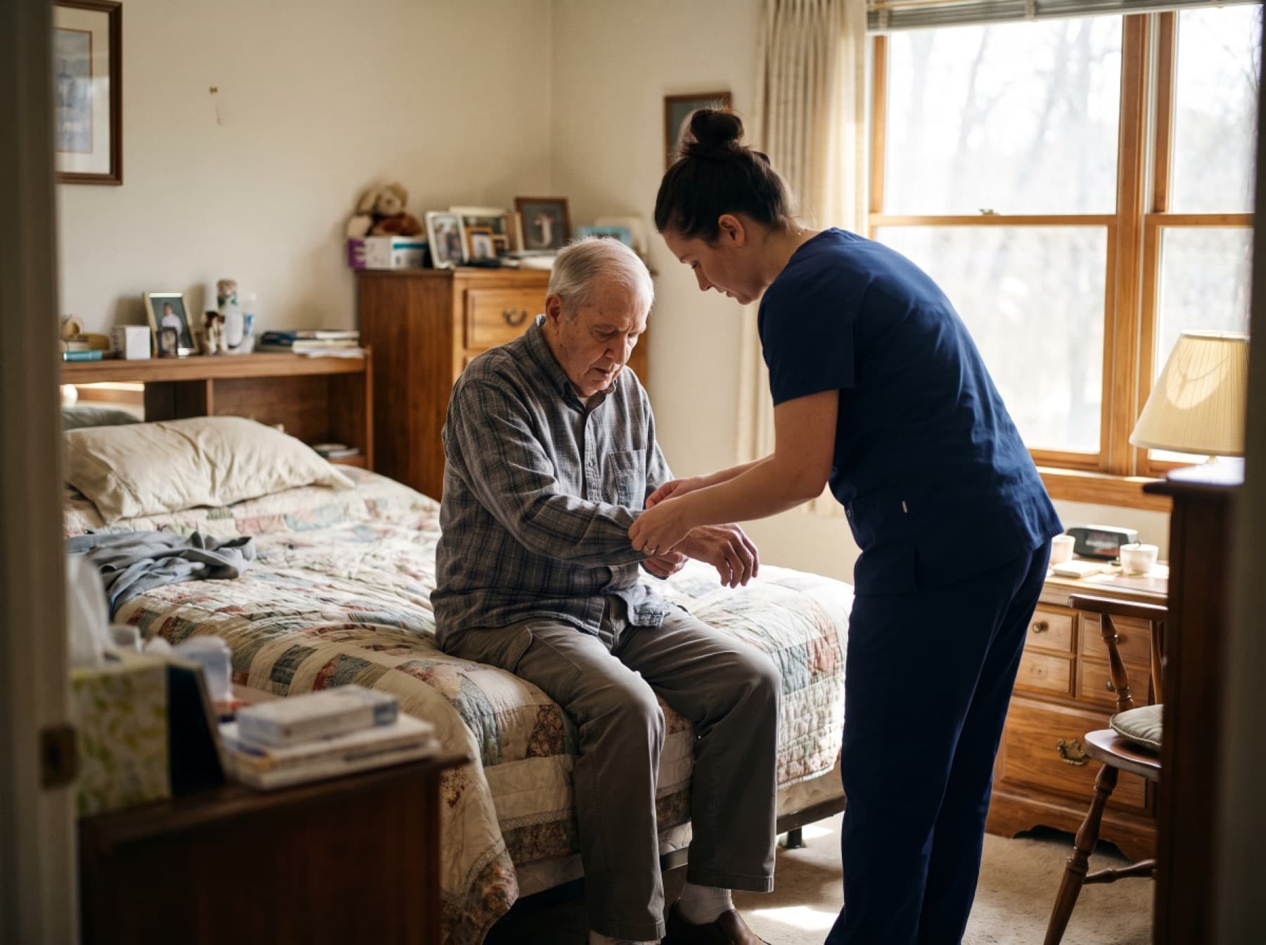 A caregiver in navy scrubs helping an older man fasten his shirt cuff as he sits on the edge of his bed.