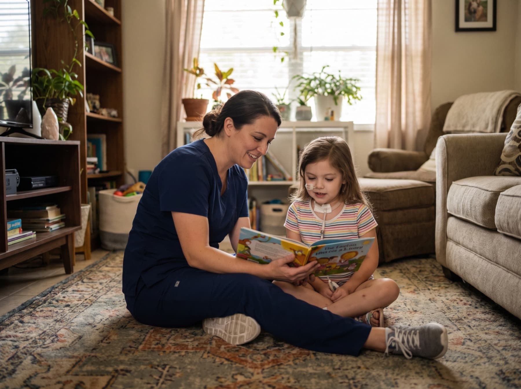 A caregiver in navy scrubs sitting on the floor reading a picture book with a young girl, both absorbed in the story.